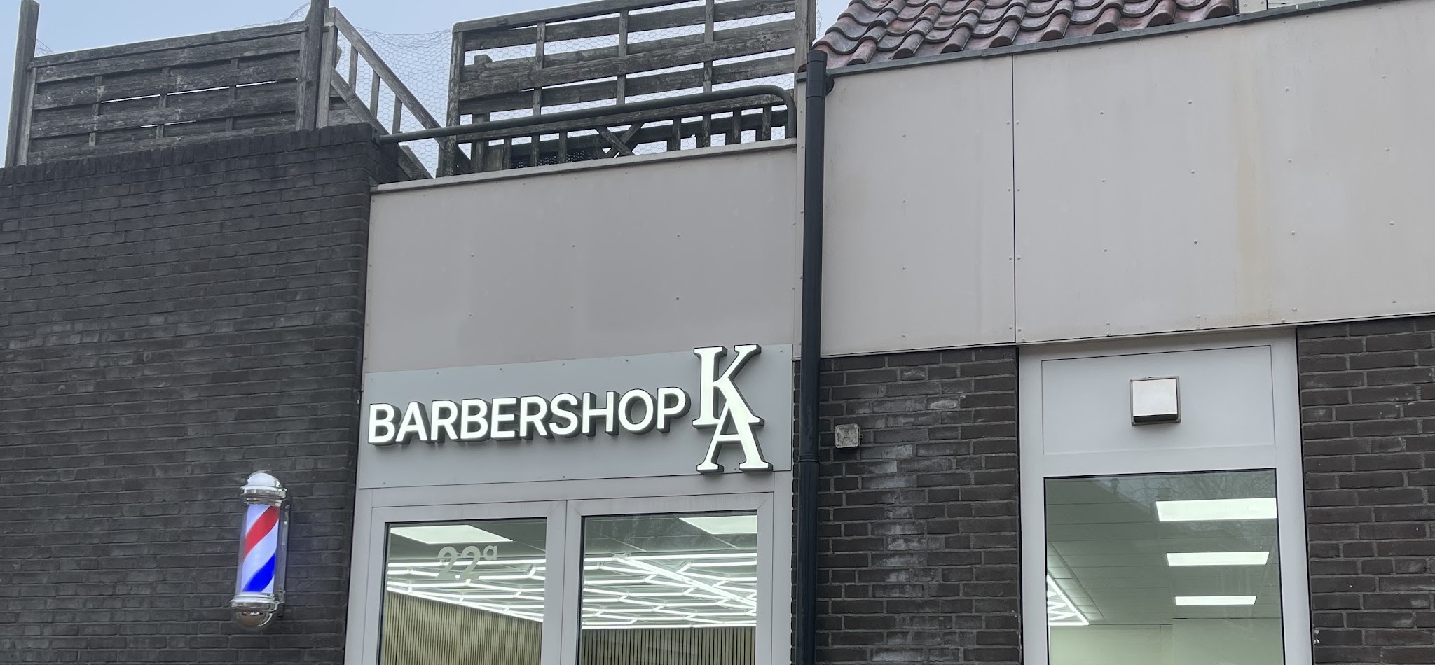 De gevel aan de Dorpsstraat overdag: bord BARBERSHOP KA boven de glazen deuren en de kapperspaal aan de muur