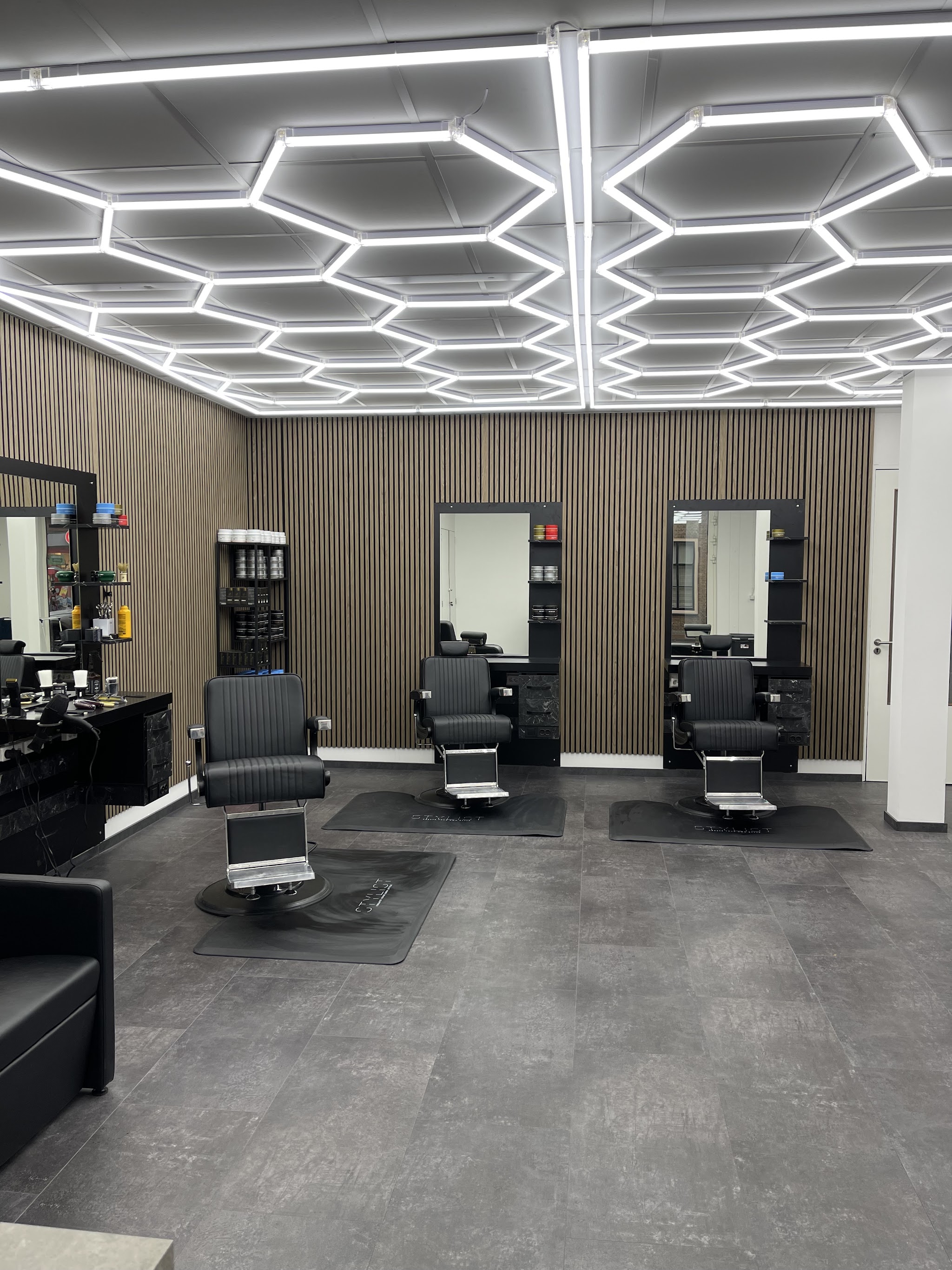 De zaak van Barbershop KA: drie zwarte kappersstoelen voor spiegels, houten lamellenwanden en een plafond vol zeshoekige lichtlijnen