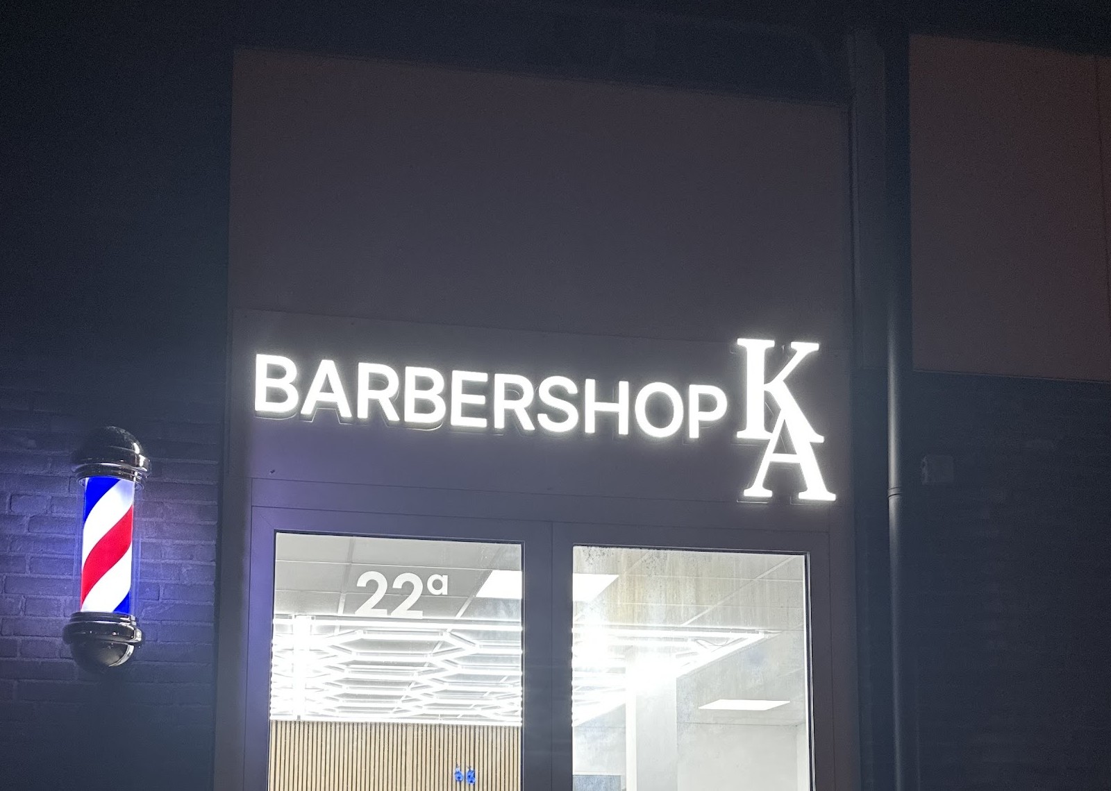 De gevel van Barbershop KA in het donker: verlicht bord BARBERSHOP KA, de draaiende kapperspaal en de verlichte zaak achter de deur